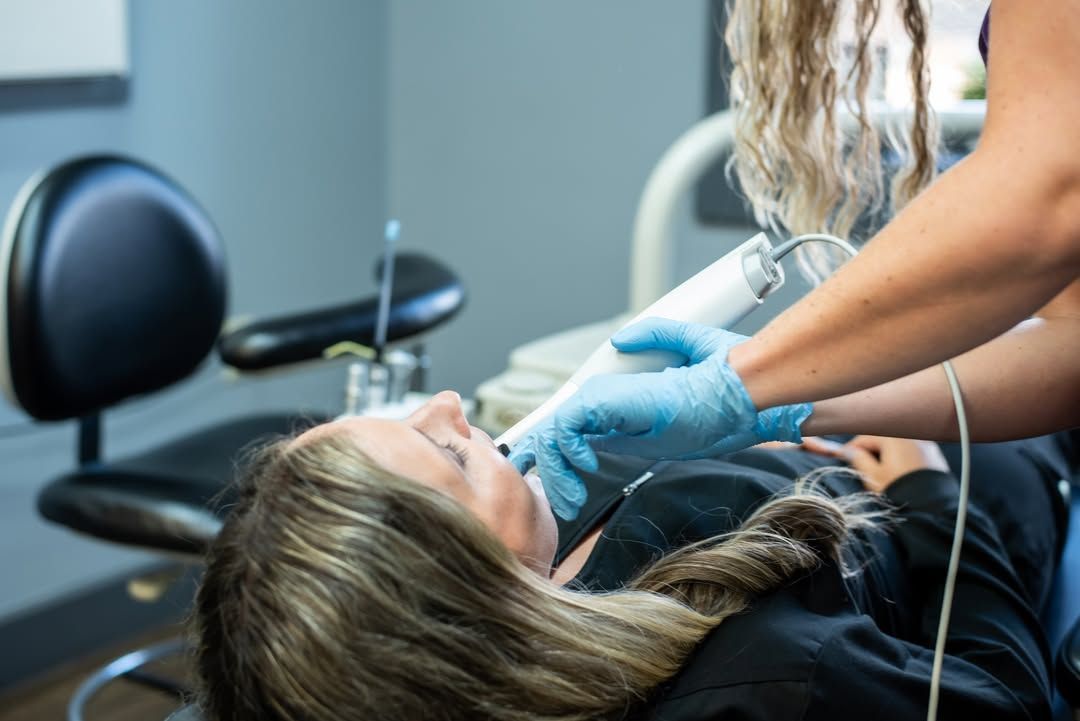 Woman in dentist chair; dentist in blue gloves using a dental instrument, grey wall.