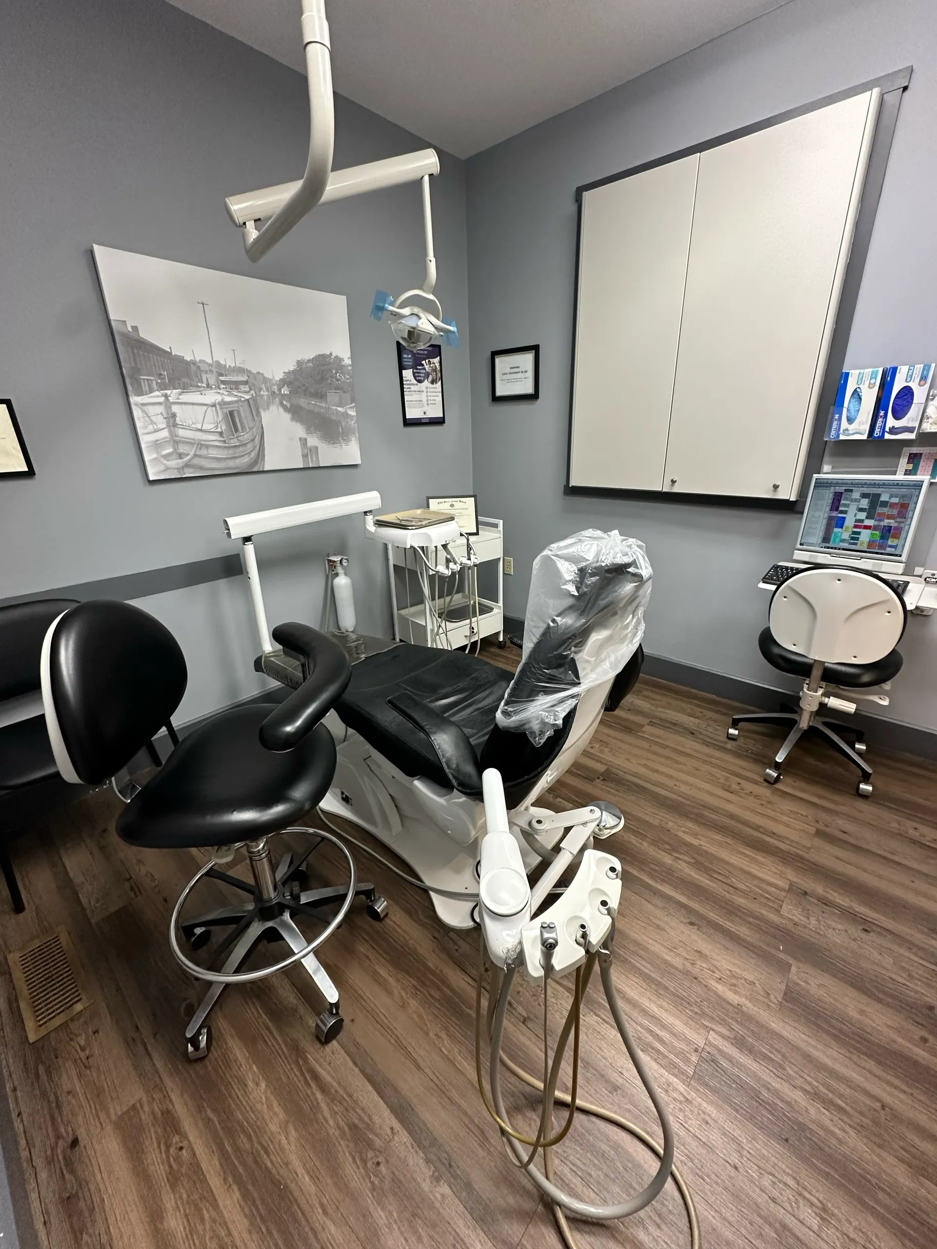 Dental office with chair, equipment, and grey walls. Artwork and a large white board are visible.