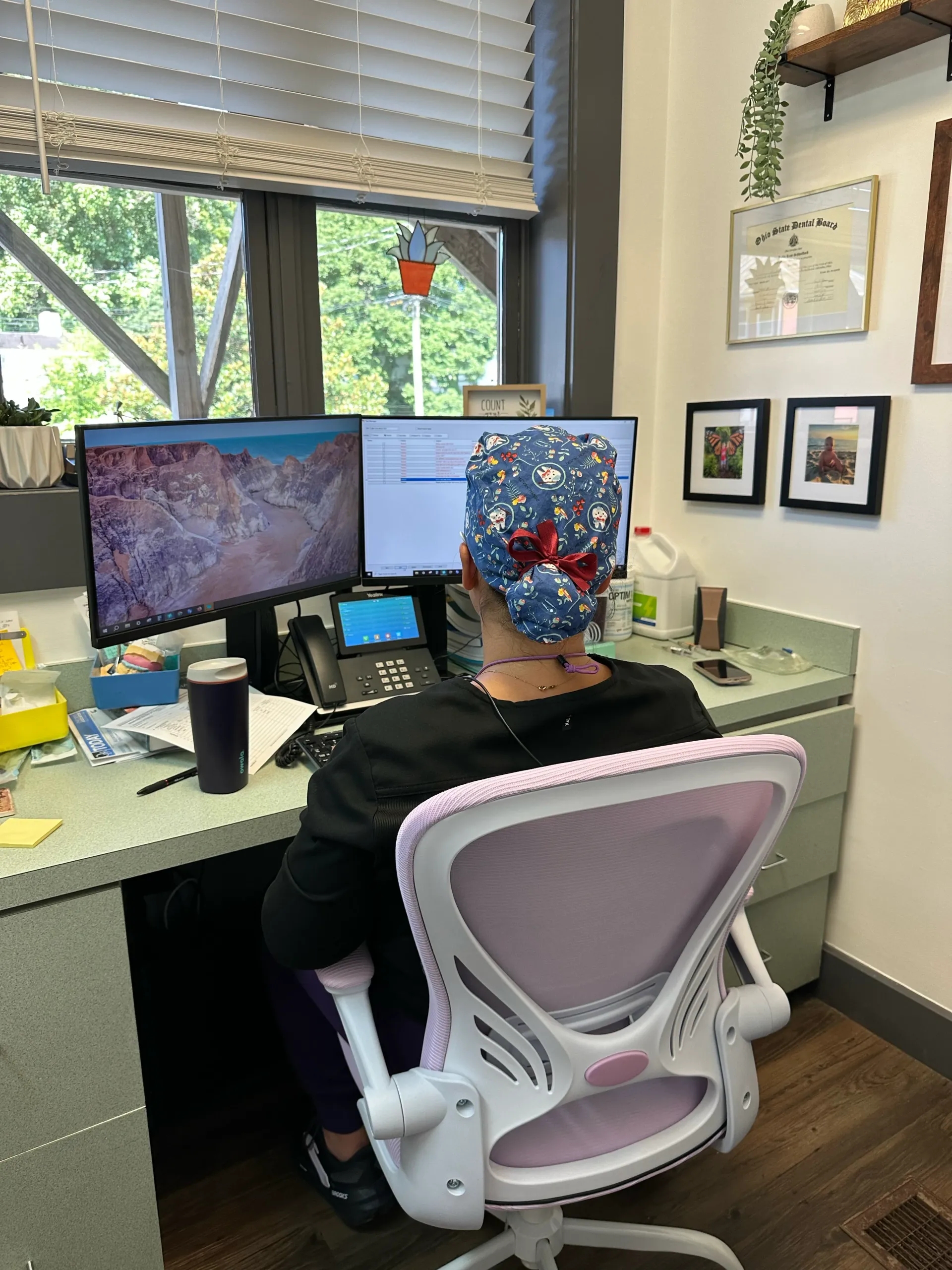 Person wearing a floral scrub cap, seated at a desk with dual monitors, working in a brightly lit office.