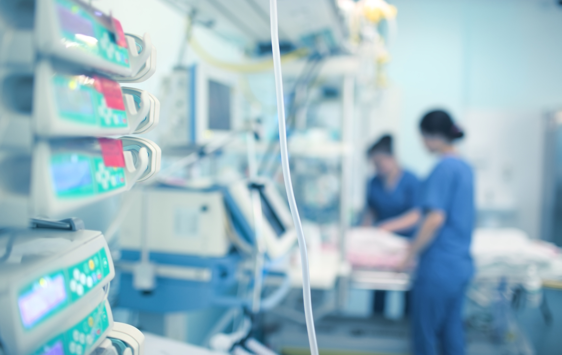 Medical monitors and equipment in a hospital room; two healthcare workers in scrubs are blurred in the background.