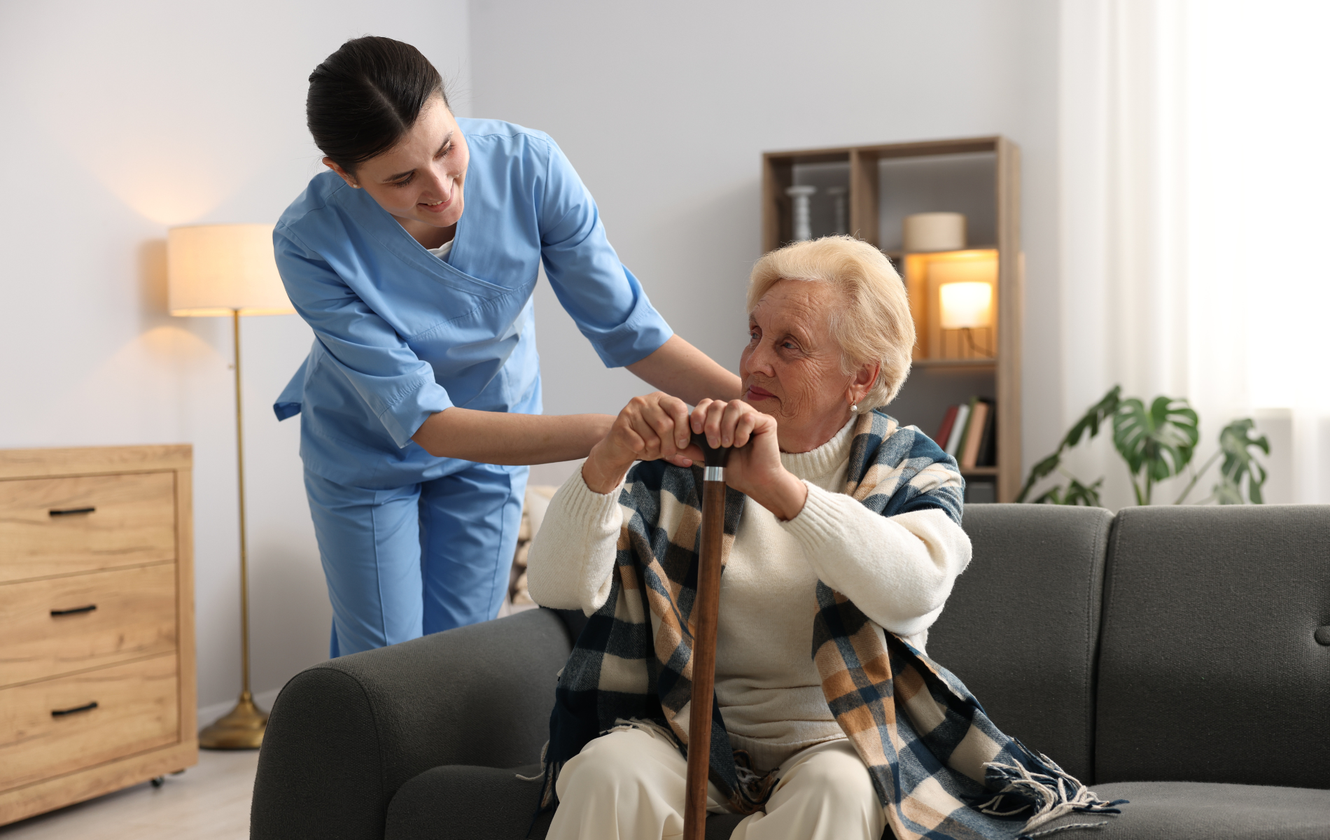 Caregiver assisting a senior woman with a cane sitting on a sofa.