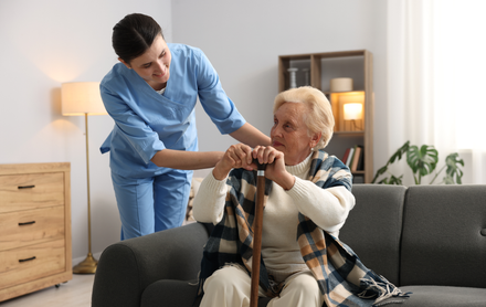 Caregiver assisting a senior woman with a cane sitting on a sofa.