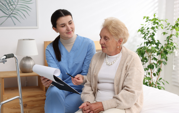 Nurse showing a clipboard to an older adult seated on a bed. Bright room with walker.