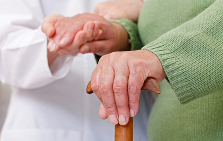 Person's hand on a cane, held by someone in a white coat and green sweater for support.