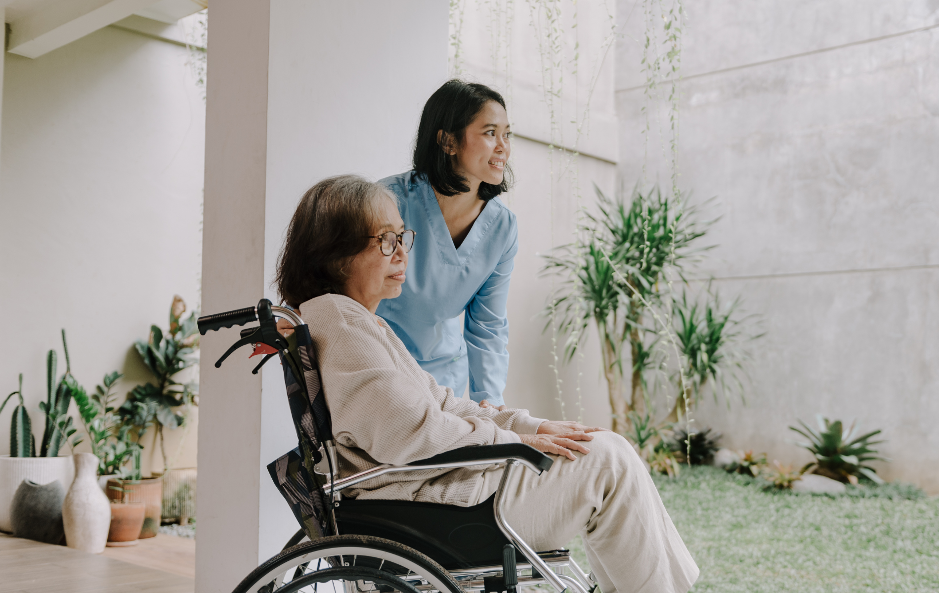An older adult uses a cane with assistance from a caregiver in a well-lit living room.