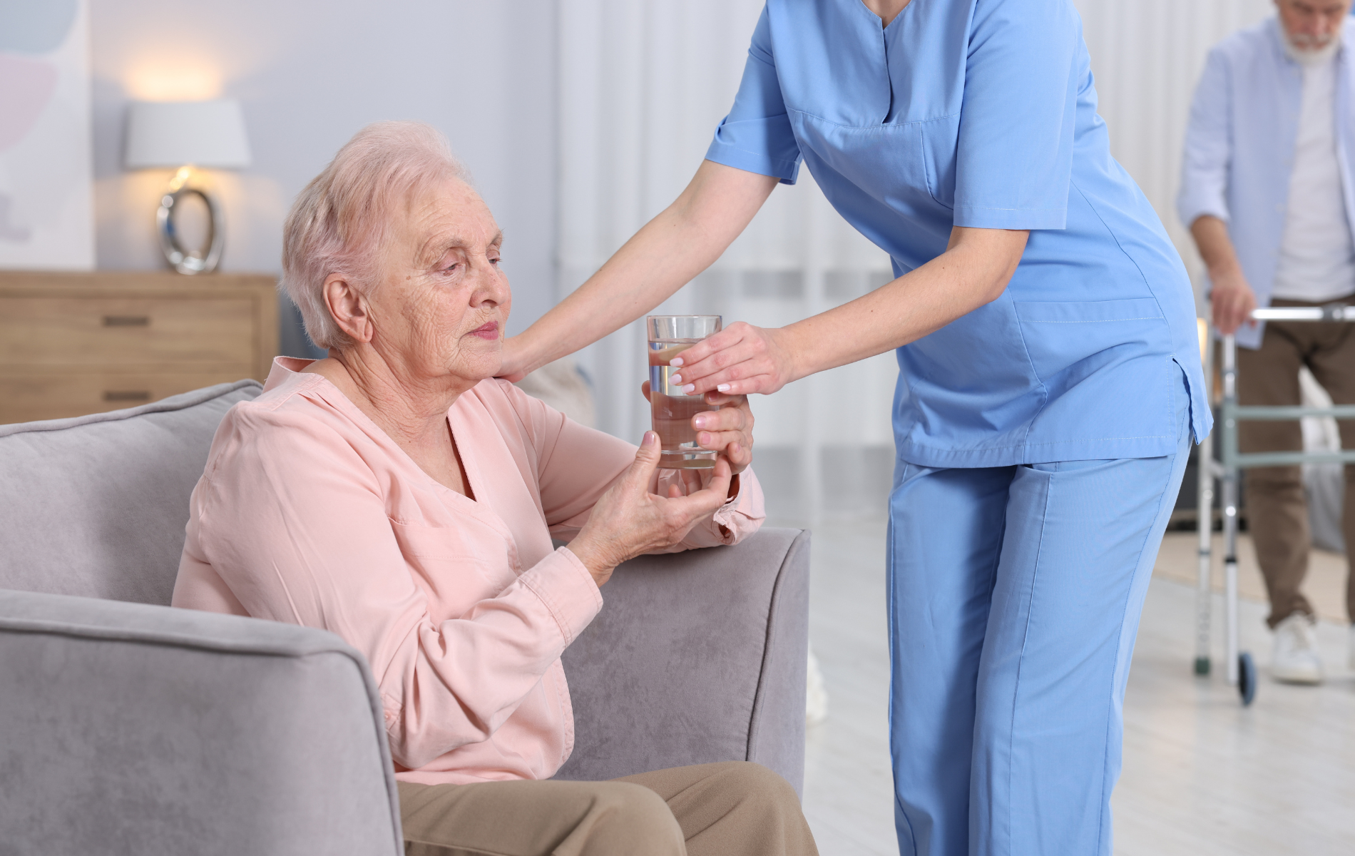 A caregiver in blue scrubs offers water to an elderly person seated in a gray chair.