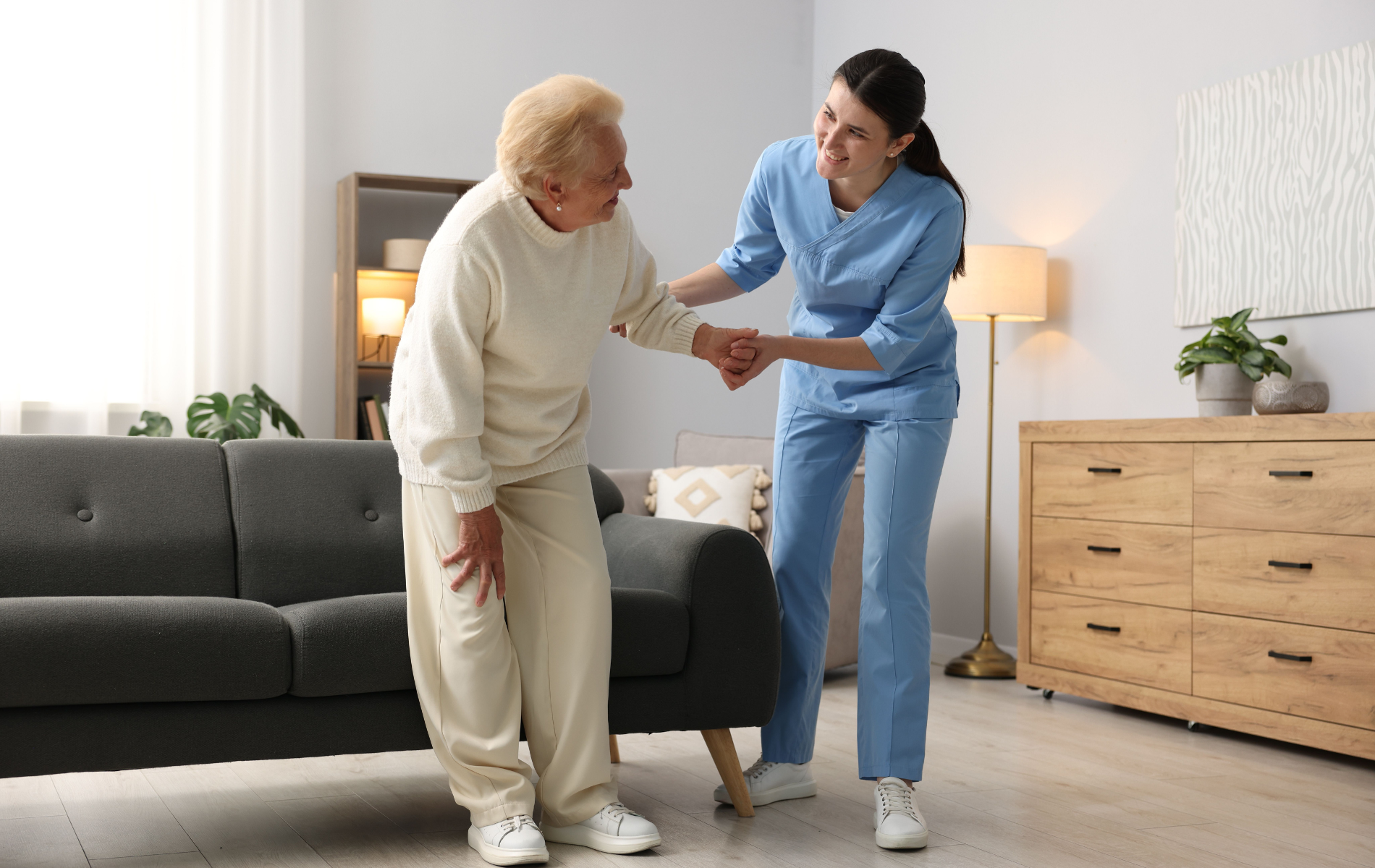 Caregiver assisting a person in a living room; caregiver in blue uniform helps person in sweat suit to stand.