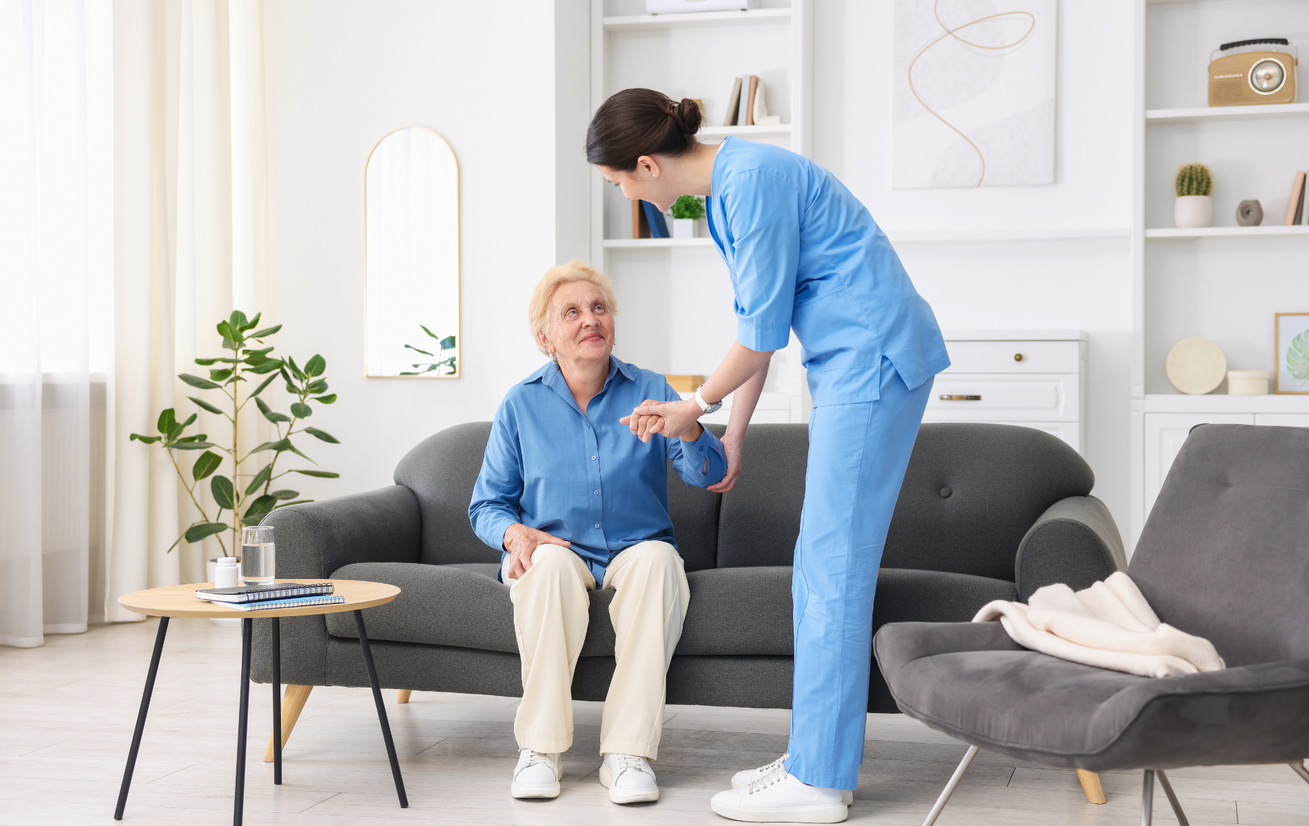 Nurse assists a person from a sofa in a bright living room, checking their wrist.