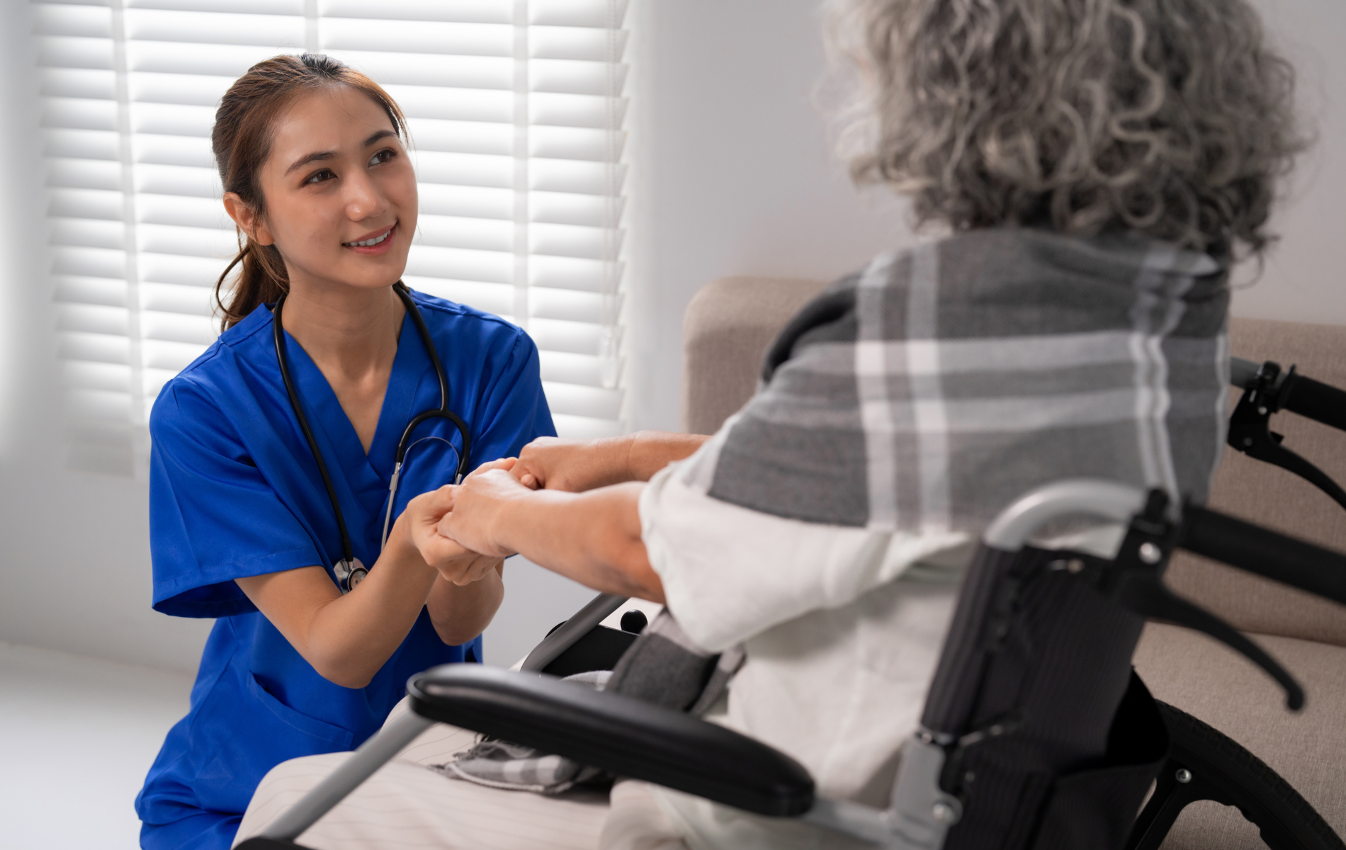 Caregiver holding hands with a person in a wheelchair indoors; caregiver smiling.