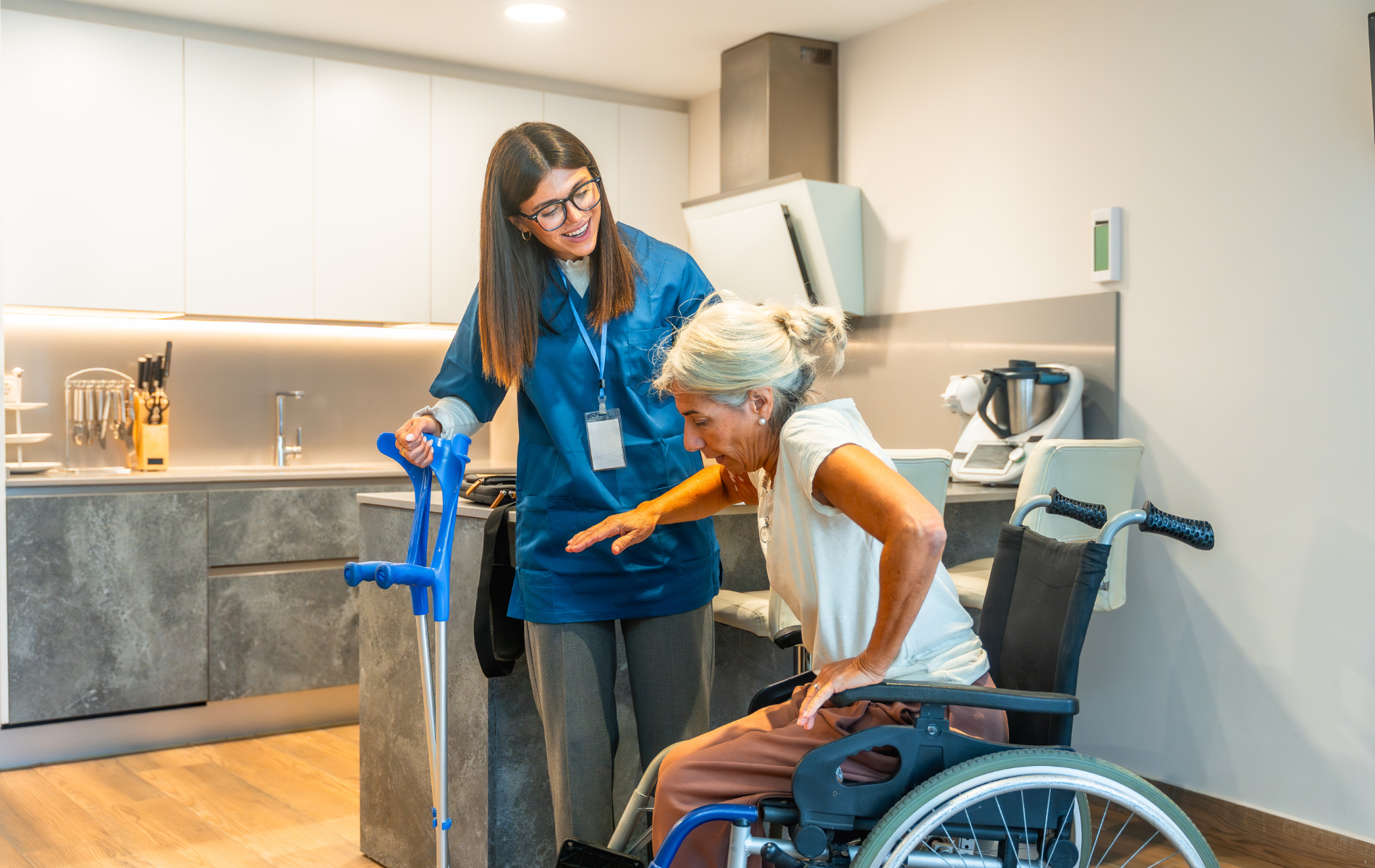 A caregiver assisting a person with mobility issues to stand from a wheelchair in a kitchen, crutches nearby.