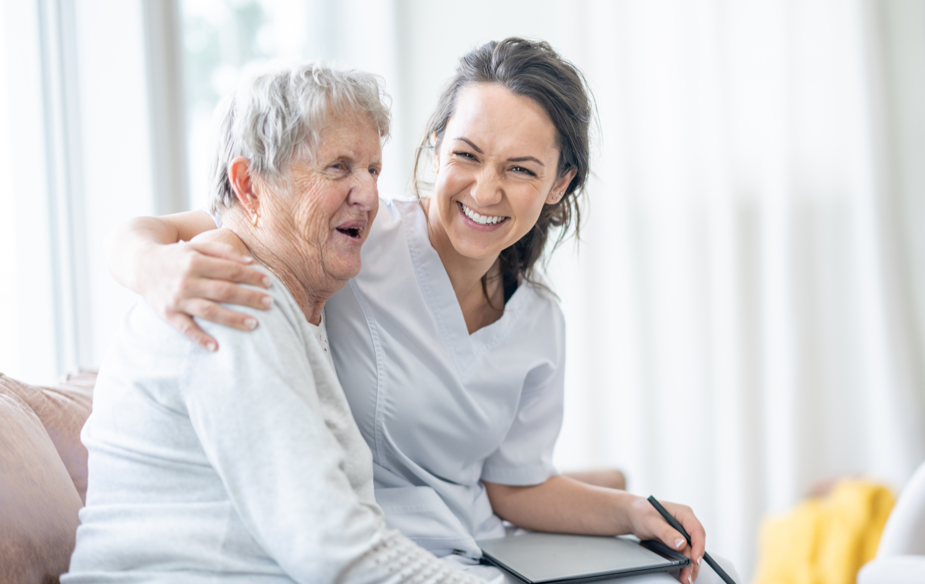 Woman in scrubs hugs and smiles at an older adult seated on a couch, indoors.