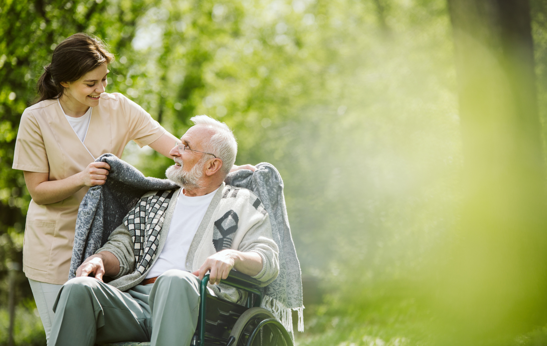Caregiver assisting a person in a wheelchair with a blanket outdoors; green setting.