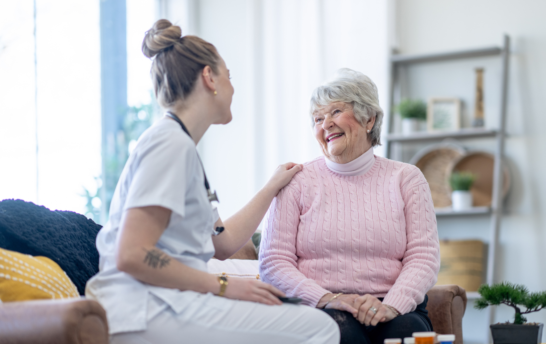 Healthcare worker comforting an older adult. Woman in pink sweater smiles. Indoors, warm lighting.