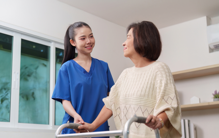 Caregiver assists elderly person using a walker in a room; both smile.