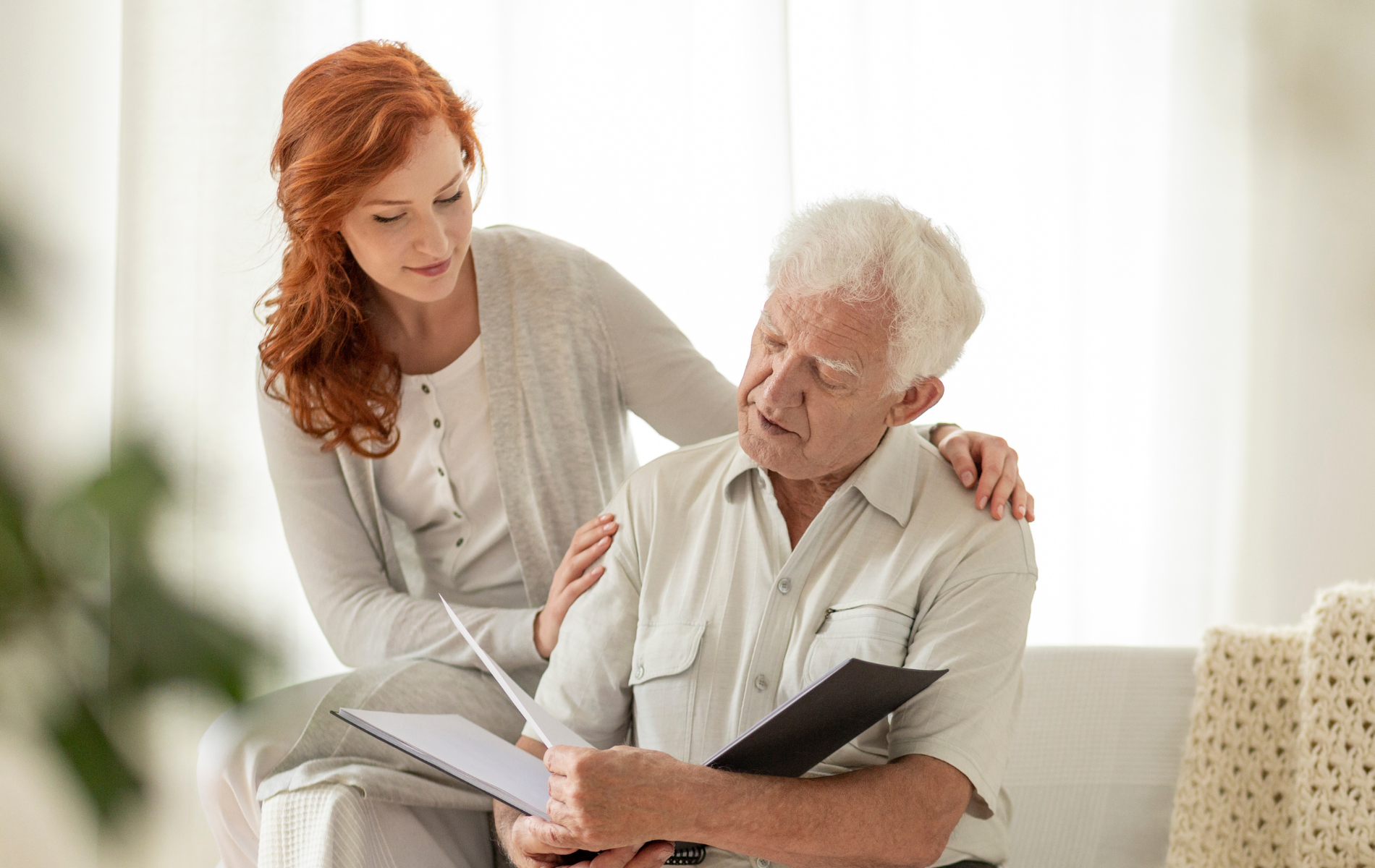 Woman with red hair comforts older man, looking at a document; indoors, soft light.