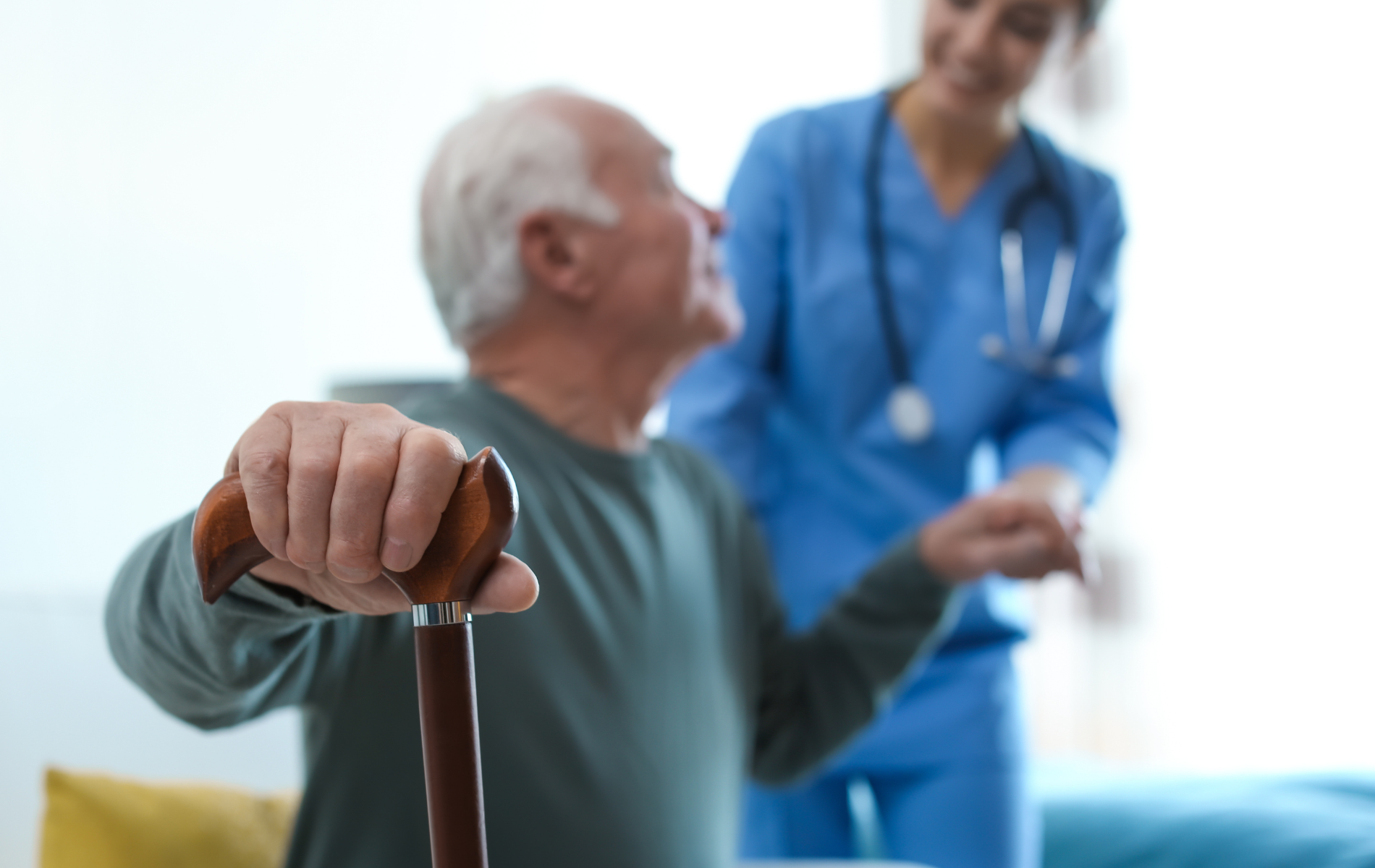 Elderly man with cane, looking at healthcare worker in blue scrubs, indoors.