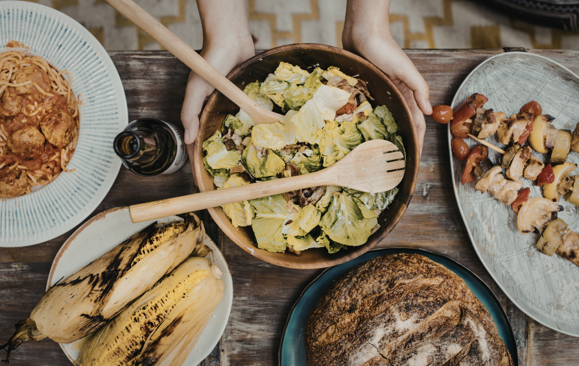 Wooden table set with a salad bowl being held, alongside bread, grilled corn, a frittata, and skewers.