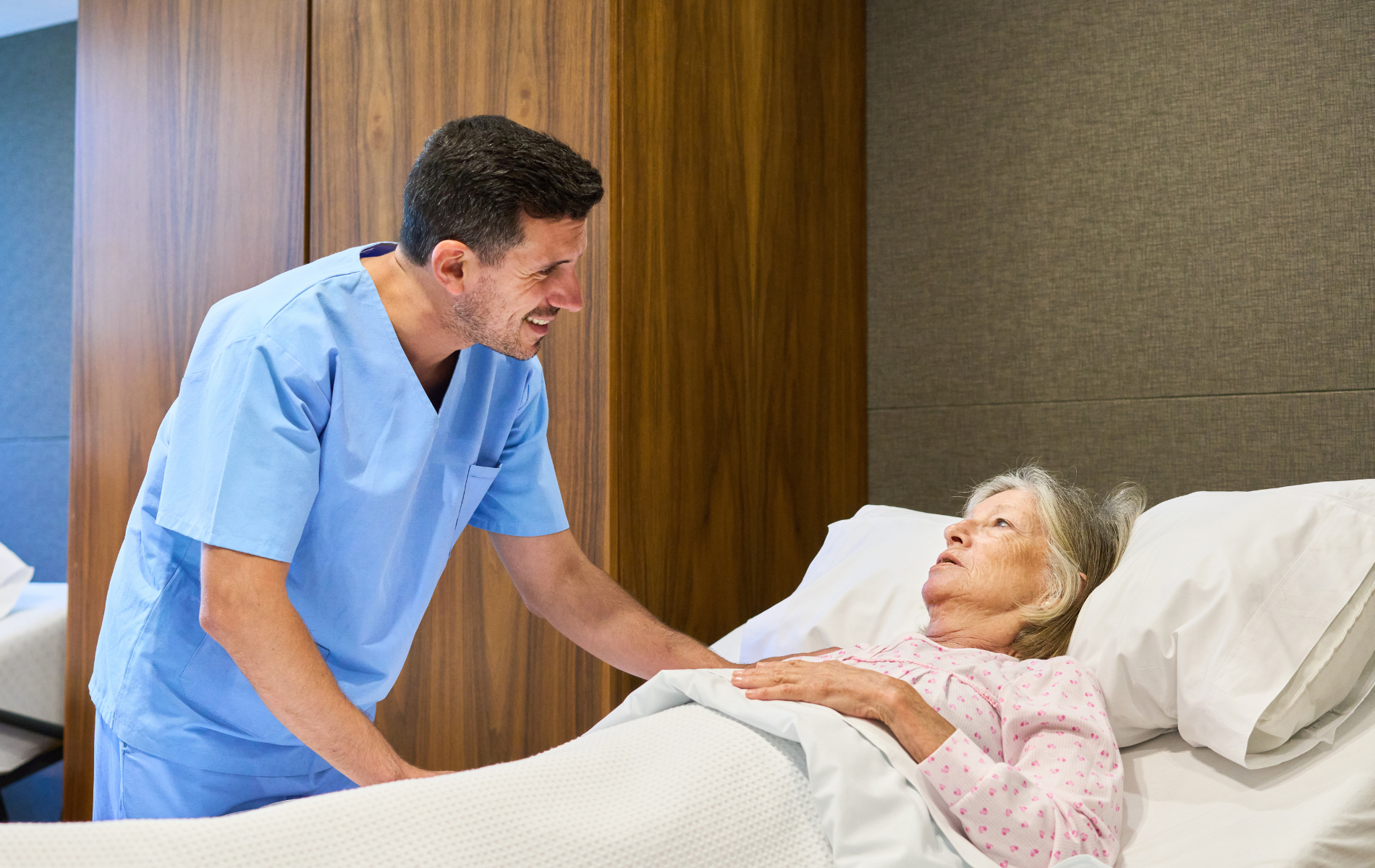 Caregiver assisting a person in a hospital bed; blue scrubs, bedside cabinet in the background.