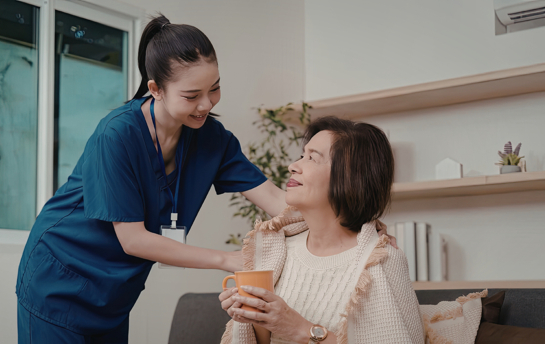 Caregiver in blue scrubs comforts elderly person with a drink. They are in a well-lit living room.