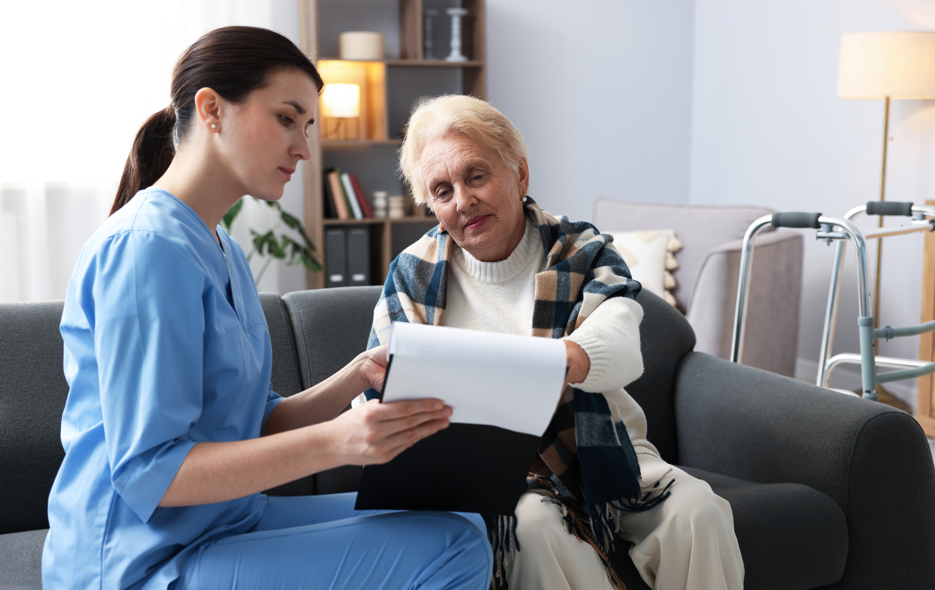 Nurse shows paperwork to an elderly person seated on a couch indoors.