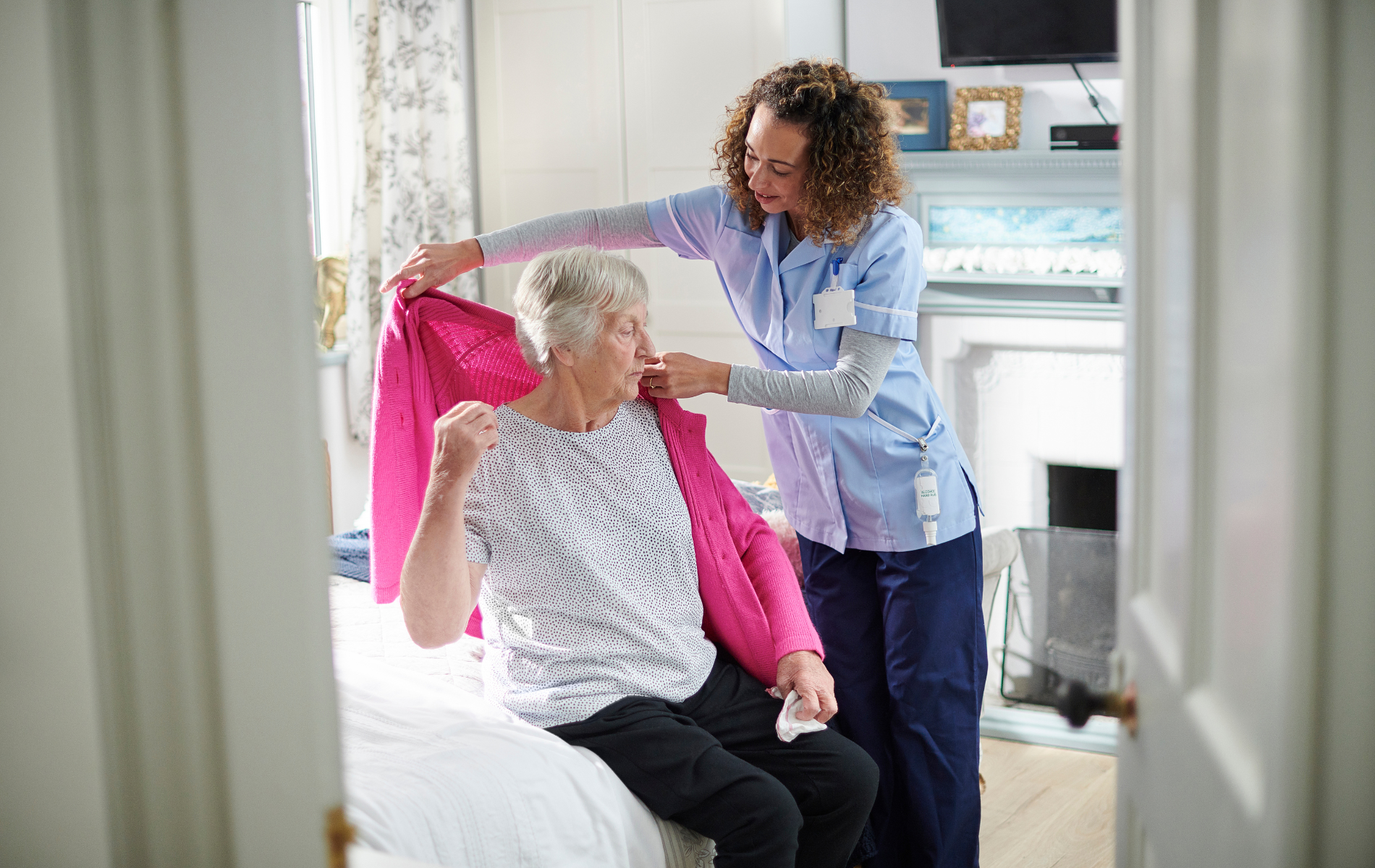 Caregiver assists a person in putting on a pink sweater in a bedroom.