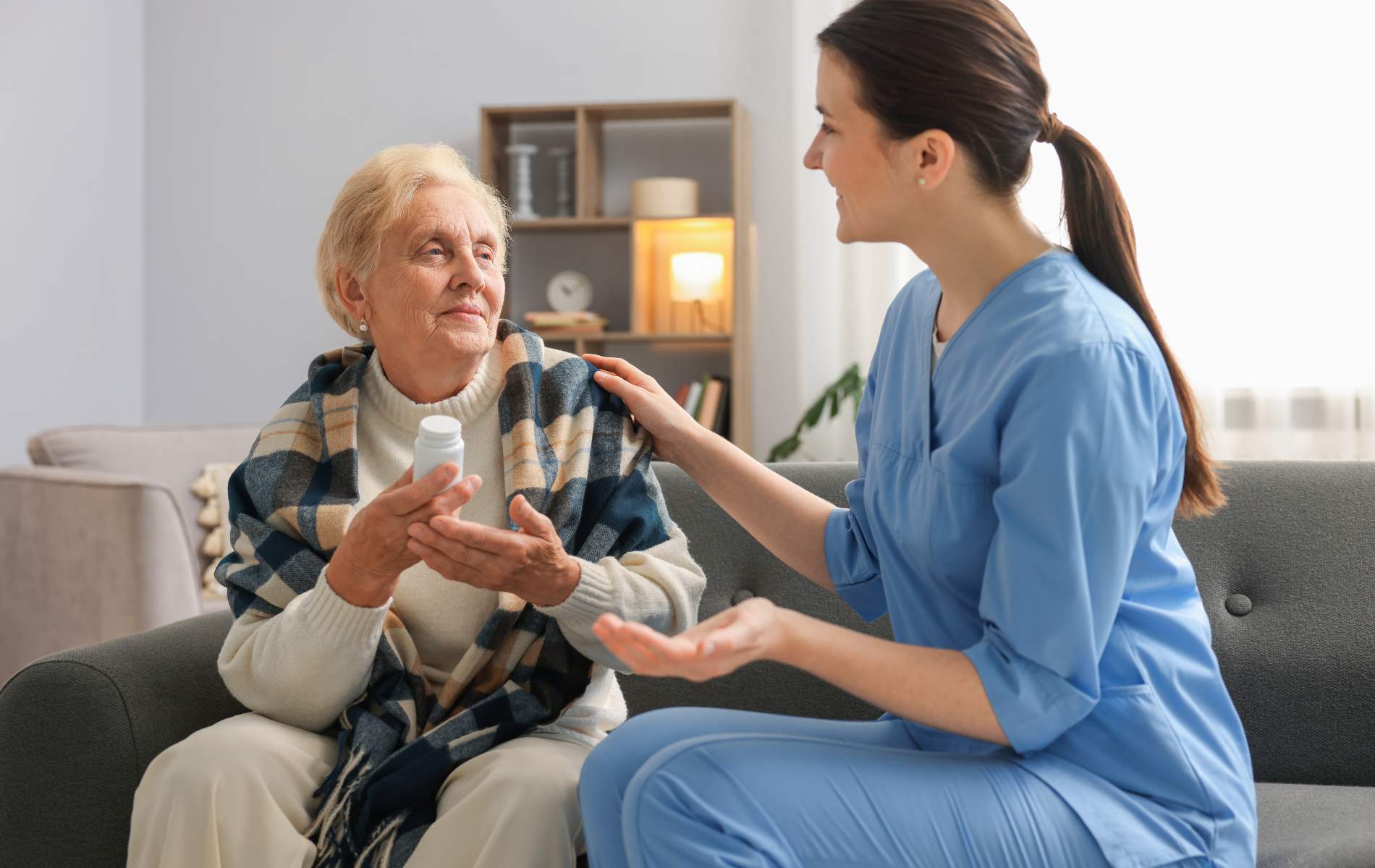 Woman in blue scrubs assists an elderly woman taking medication on a couch indoors.