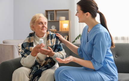 Woman in blue scrubs assists an elderly woman taking medication on a couch indoors.