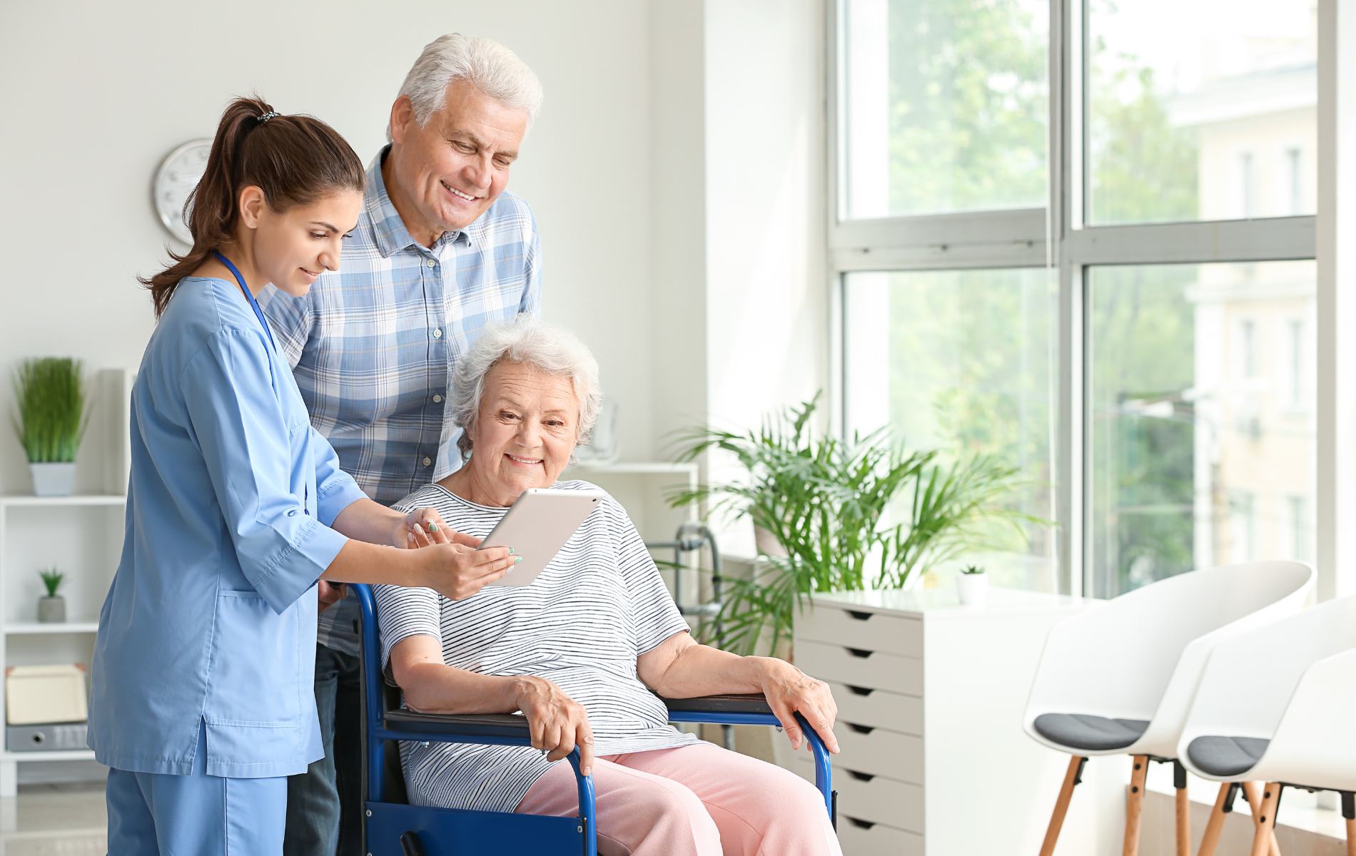 Caregiver showing tablet to an elderly woman in a wheelchair and a man; bright room.