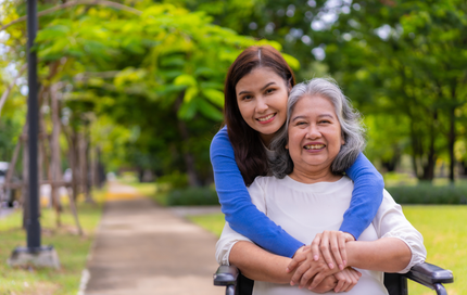 Woman hugs older woman in wheelchair outdoors, both smiling; sunny park setting.