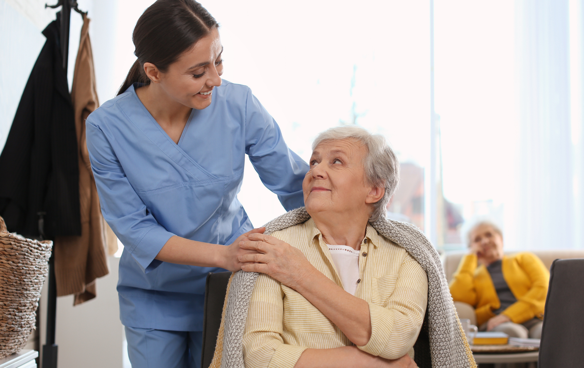 Caregiver comforts an elderly person with a blanket indoors. Another person sits in the background.