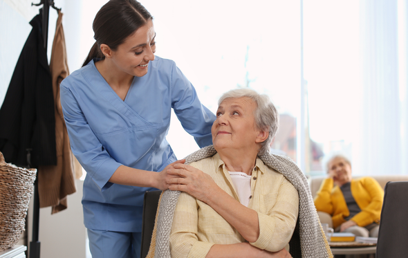 Caregiver comforts an elderly person with a blanket indoors. Another person sits in the background.