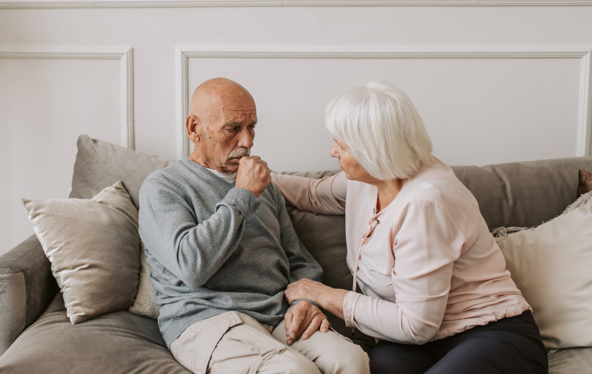 An older person coughing, comforted by a person, both seated on a couch indoors.