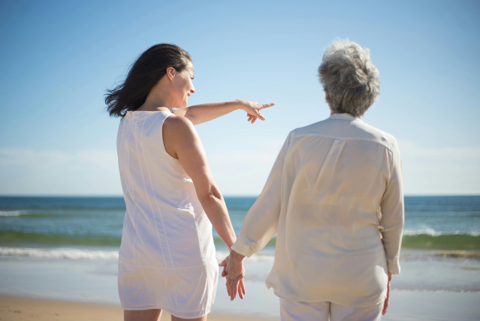 Two people holding hands on a sunny beach, with one pointing toward the ocean.