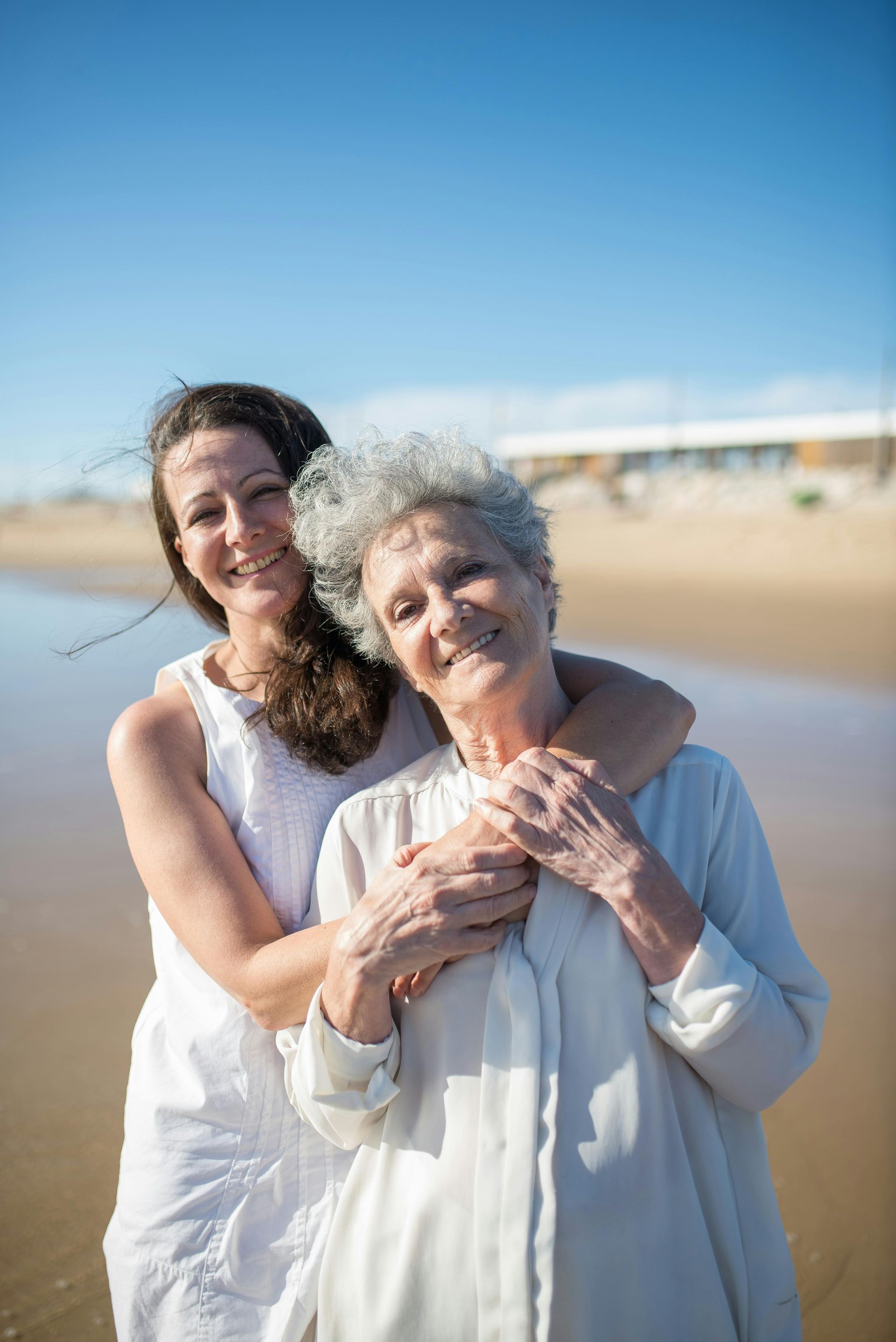 Two people embrace on a sunny beach, smiling while wearing white clothes with the ocean in the background.