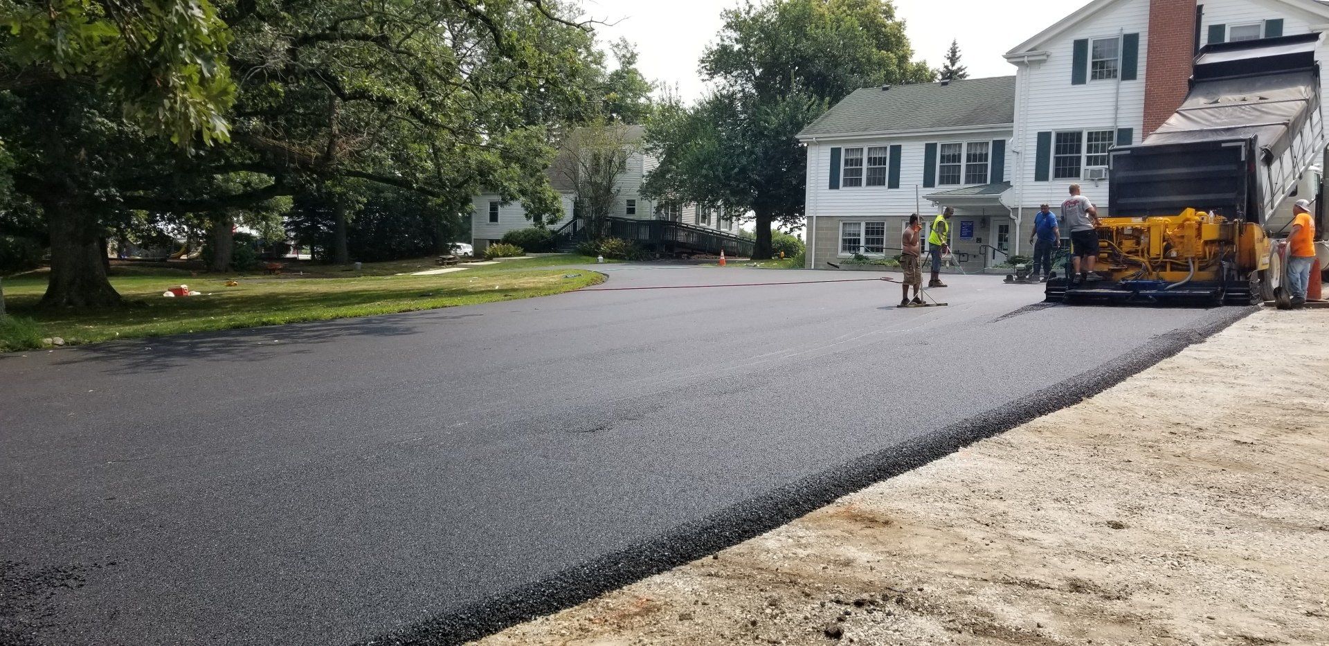 Asphalt paving a driveway with workers and equipment near a white house.