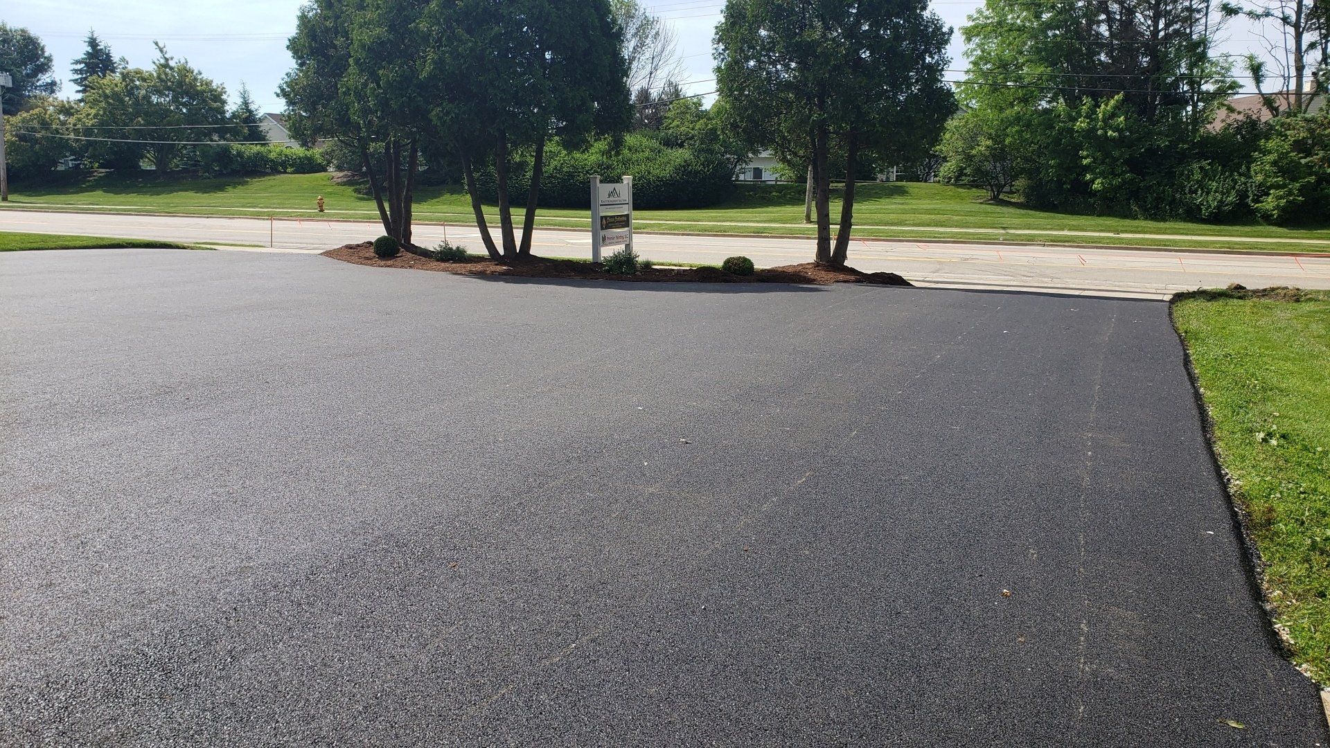Black asphalt driveway in front of a grassy lawn and three small trees.
