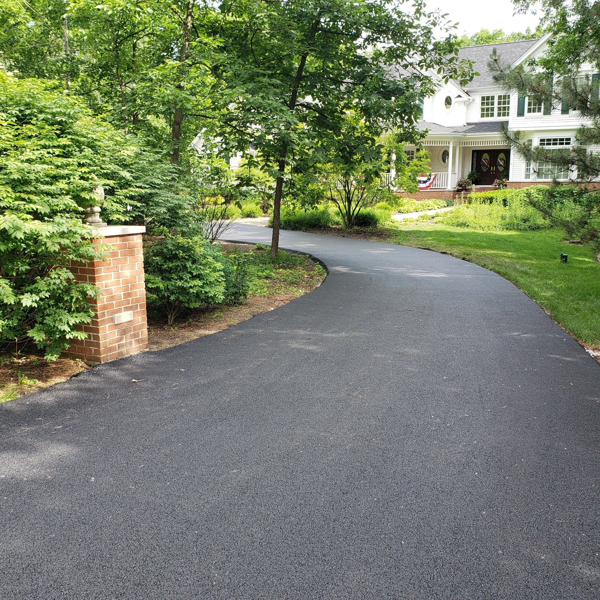 Paved driveway curves towards a white house, brick pillar on left, surrounded by trees and greenery.