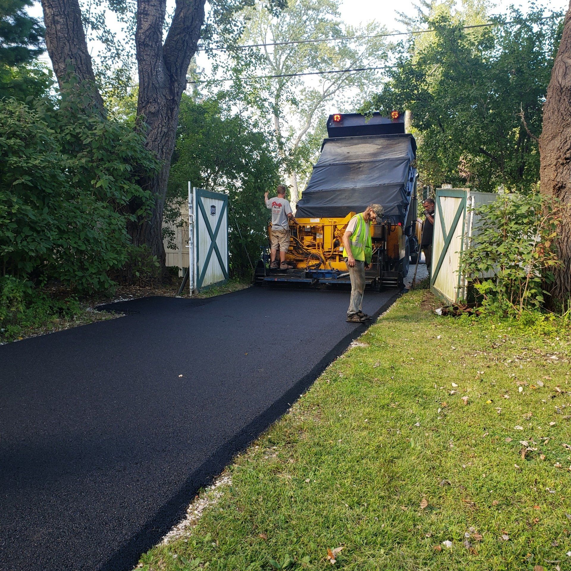Black asphalt driveway with garbage truck being loaded; two workers on back.