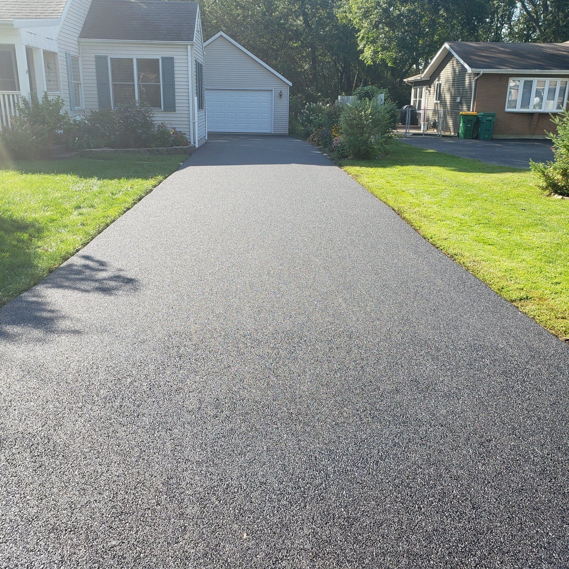 Asphalt driveway leading to a house with green grass on both sides. Sunlight is visible.