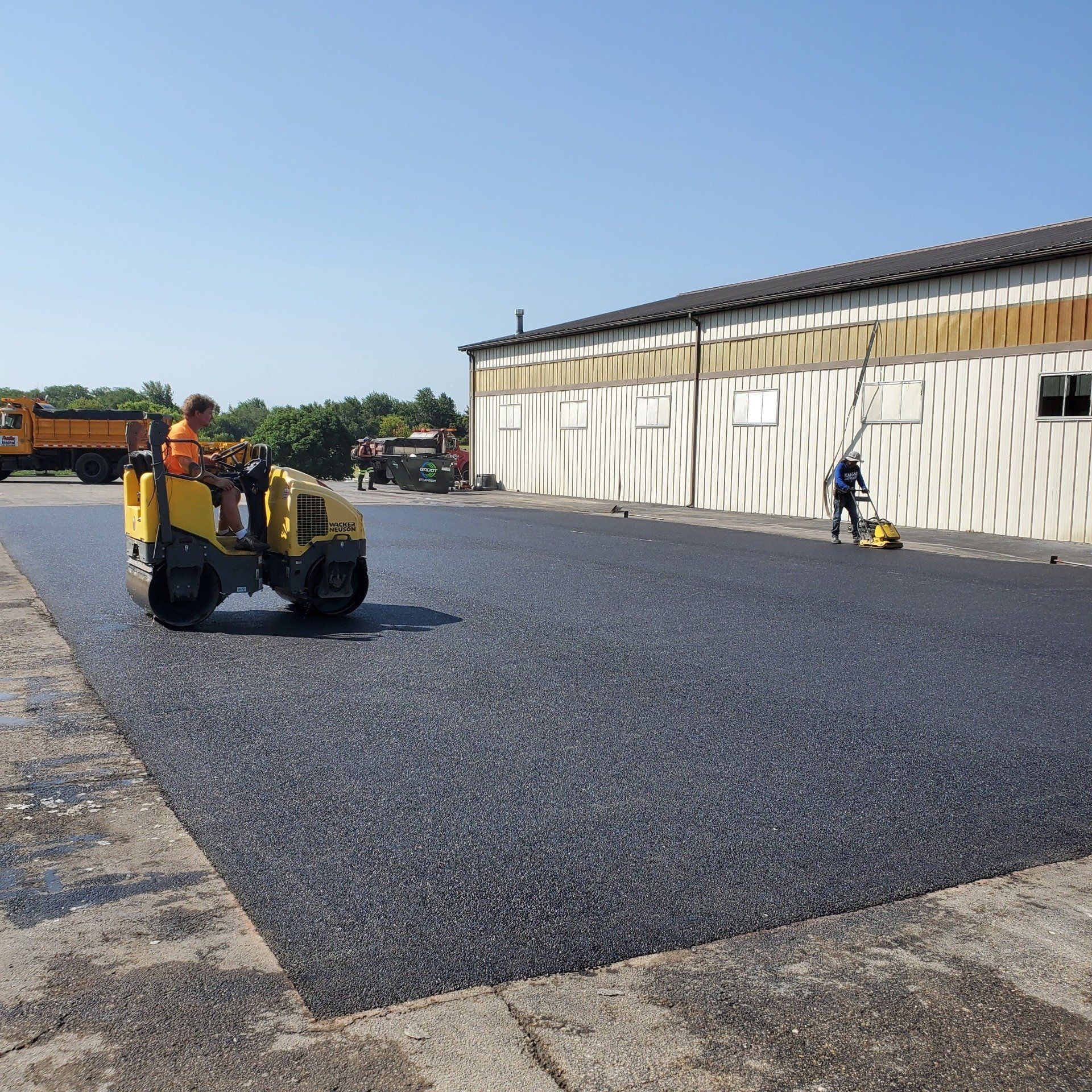 Workers paving a parking lot: a roller, a paver, a building, and a dump truck under a blue sky.