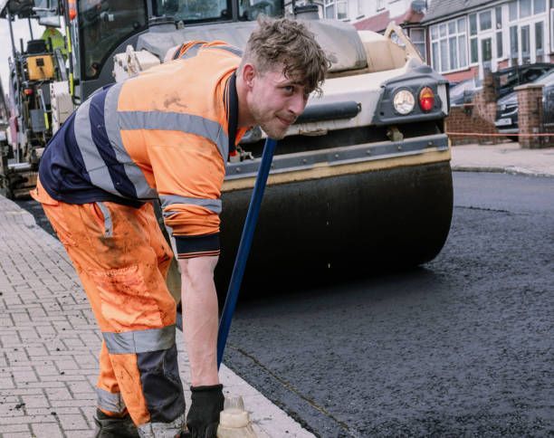 Road worker in orange and gray uniform smoothing asphalt with roller in the background.