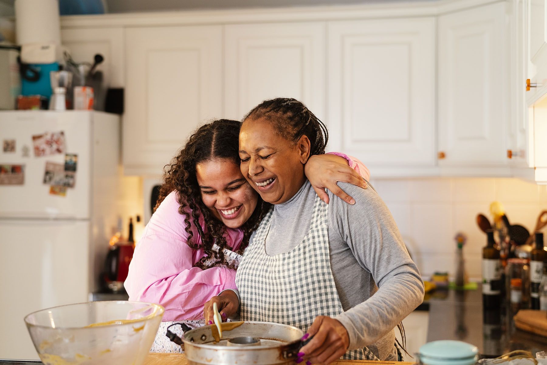 Woman and young person laughing, hugging while cooking in a kitchen.