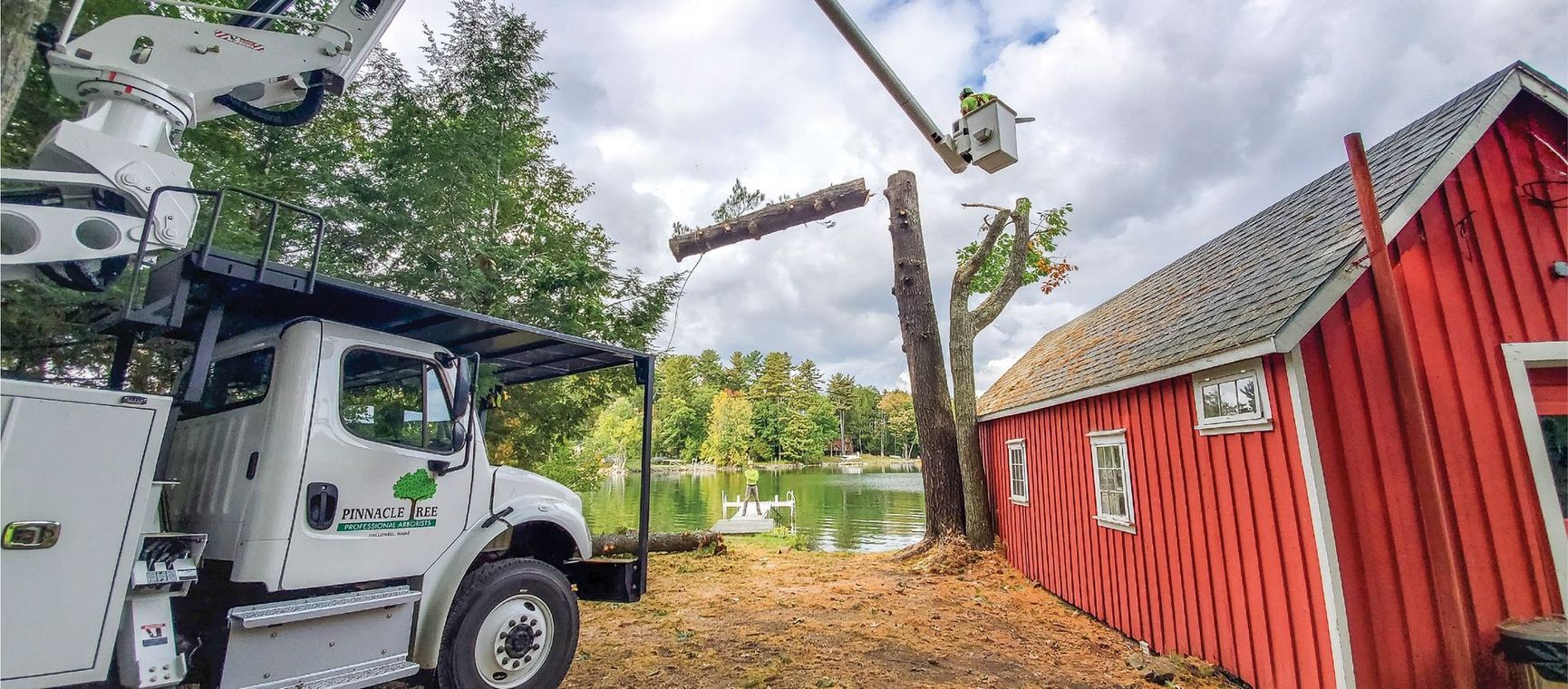 A white truck is cutting a tree in front of a red barn.