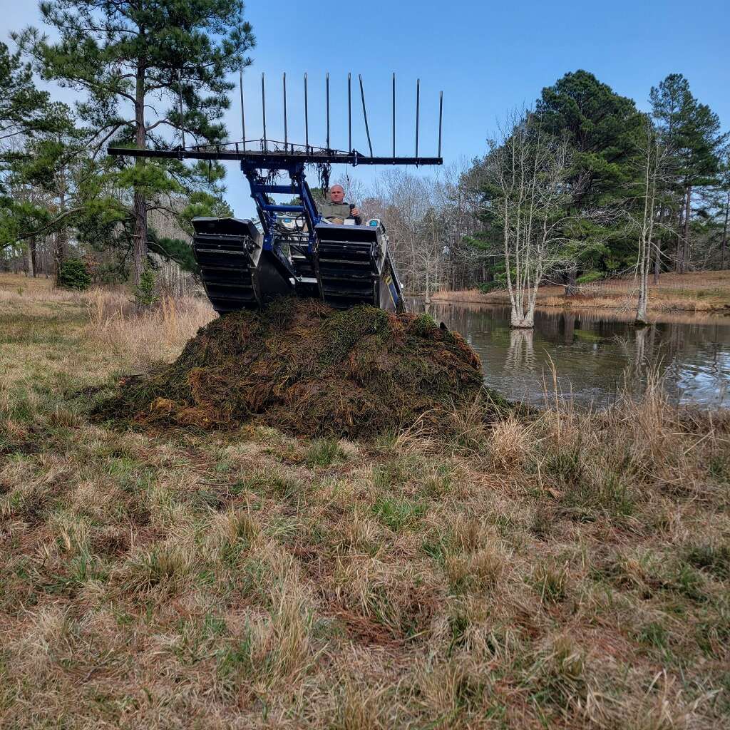 Tracked vehicle with rake harvesting pond vegetation on grassy bank.