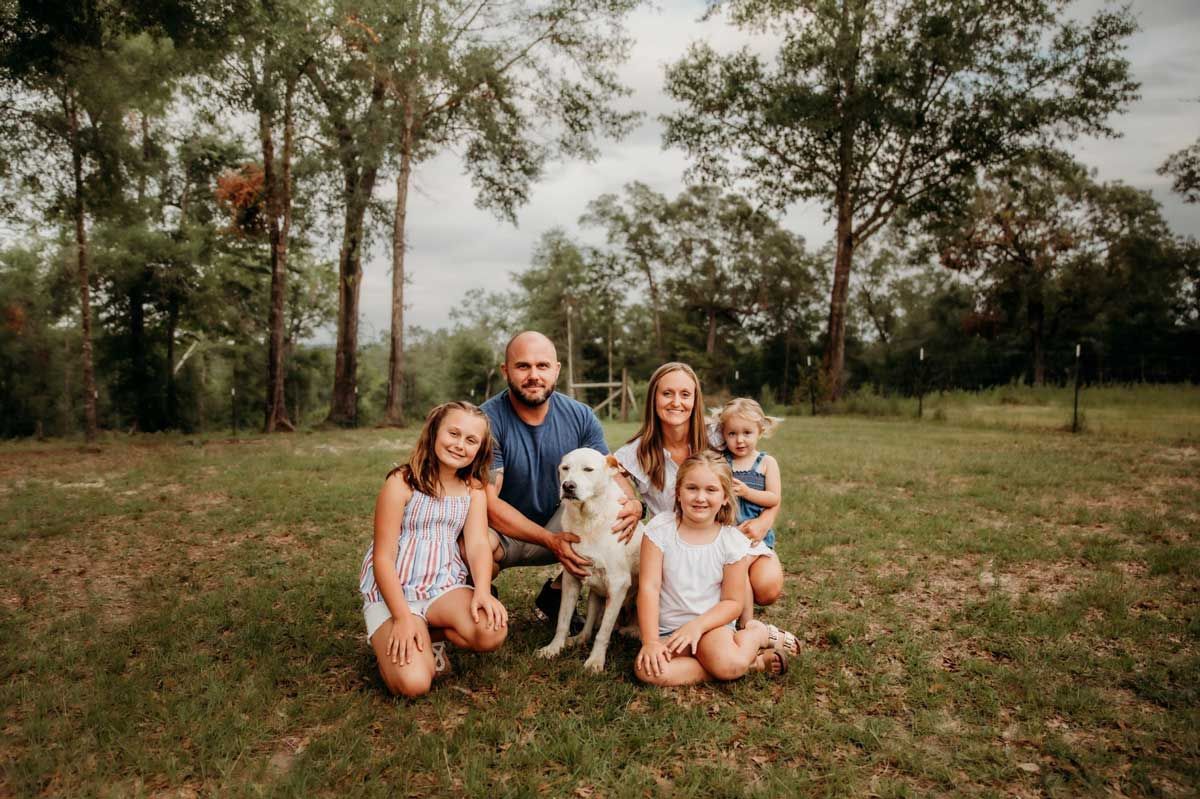 Family of five with a dog posing outdoors; they are smiling in a grassy area with trees.