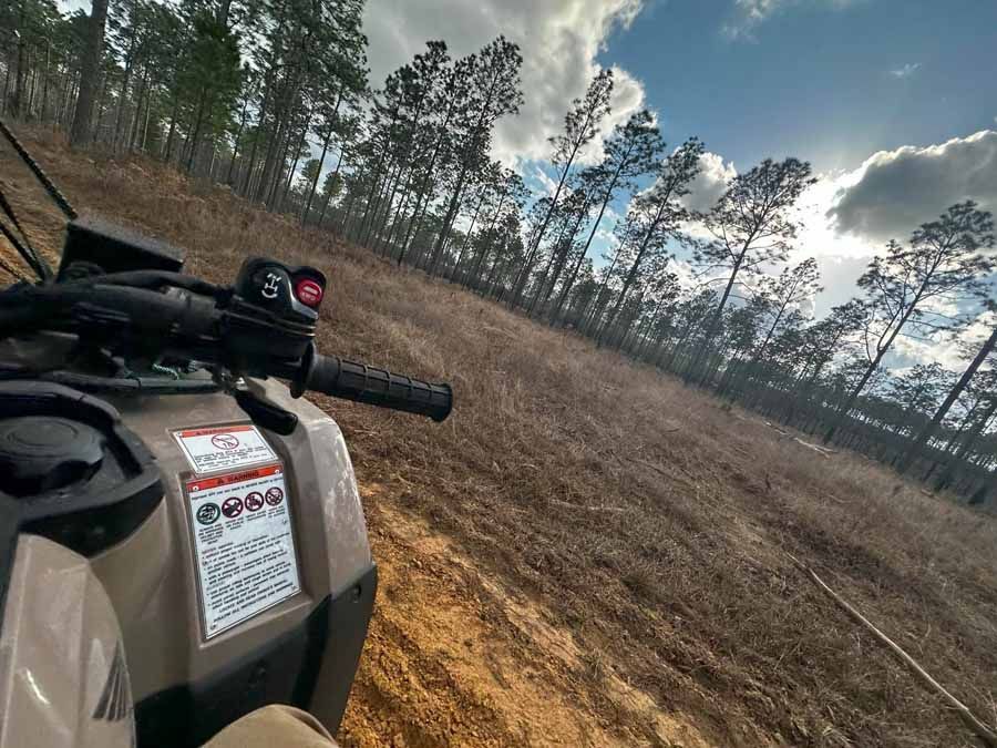 ATV on a hillside path, pine trees in the background under a partly cloudy sky.