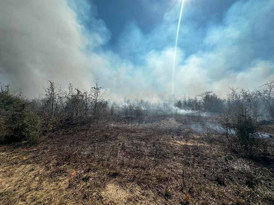 Smoke rises from a smoldering brush fire under a bright blue sky with sun streaks.