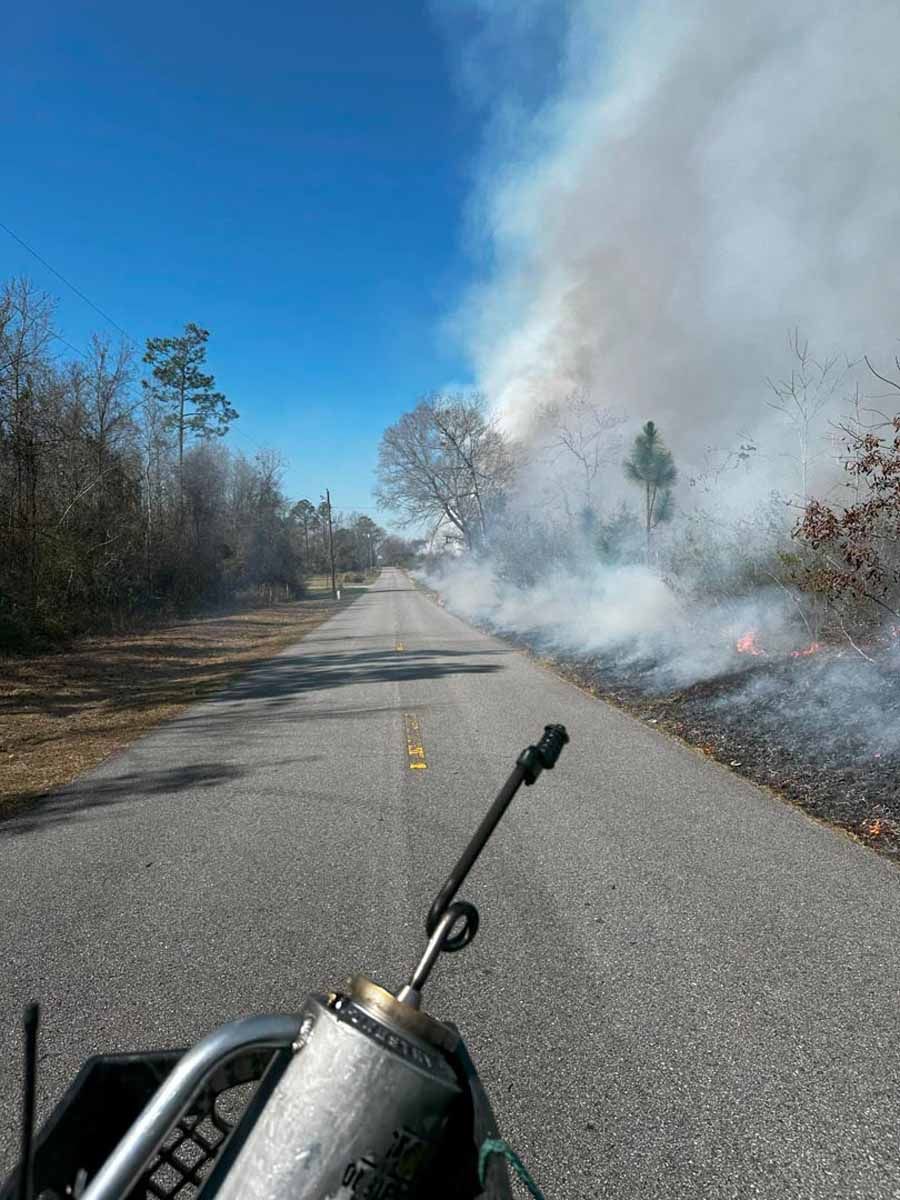 A fire burns alongside a road, smoke billowing into the blue sky. Foreground: part of a machine.
