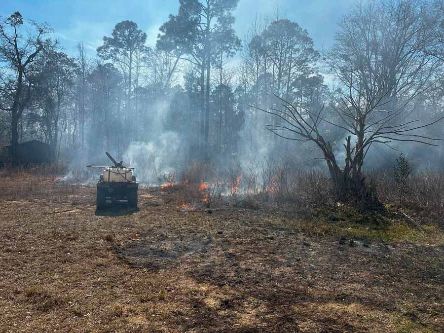 Burning brush in a field; an ATV with fire suppression equipment is present. Trees and smoke in the background.