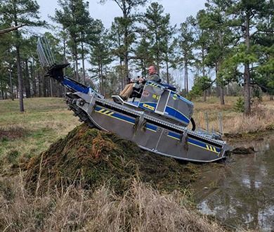 Man drives an amphibious vehicle into a pond. The vehicle is gray and blue. Outdoors.